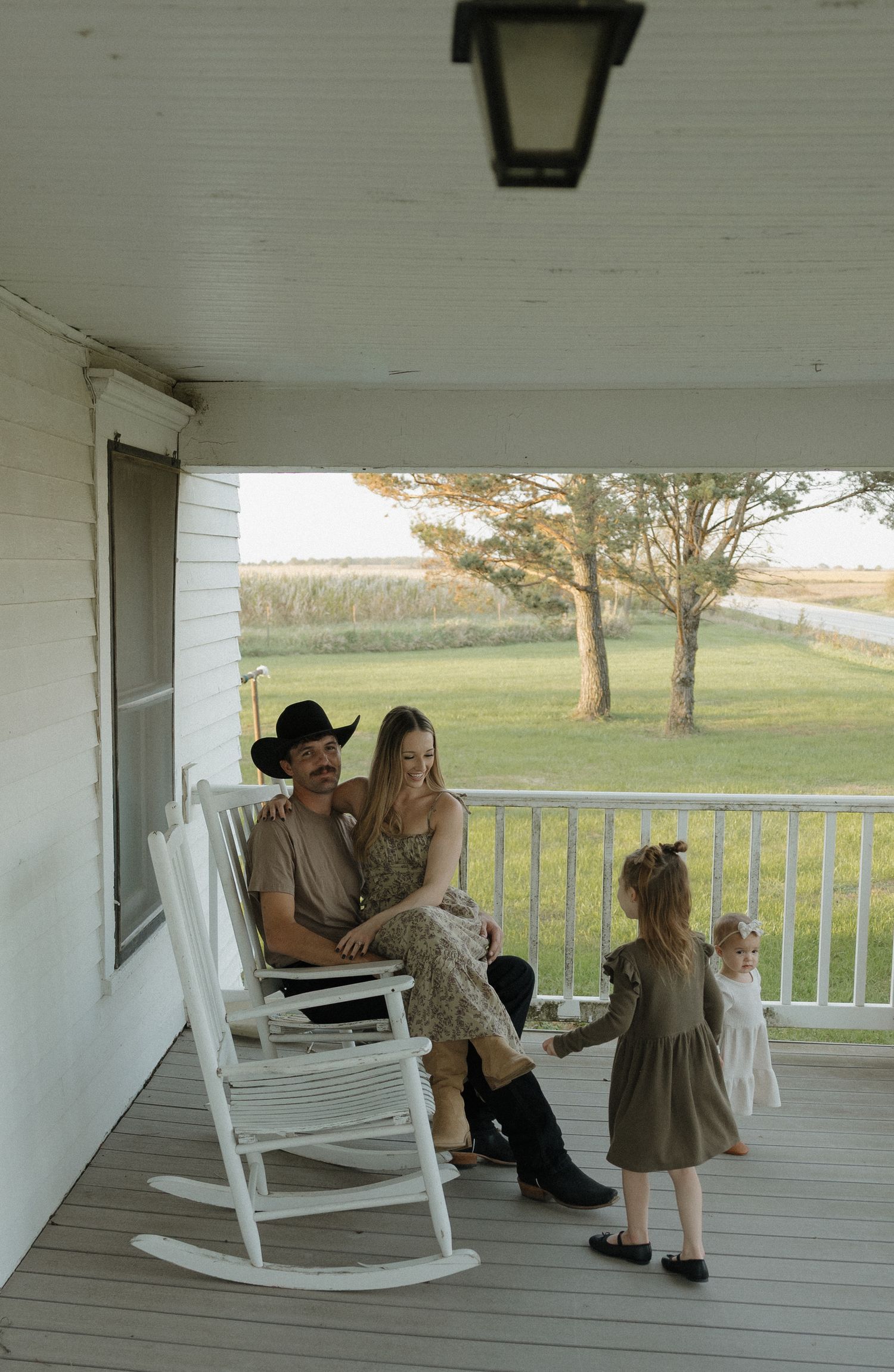 A mother in a white dress smiles while holding her baby and hugging her young daughter outdoors at golden hour.