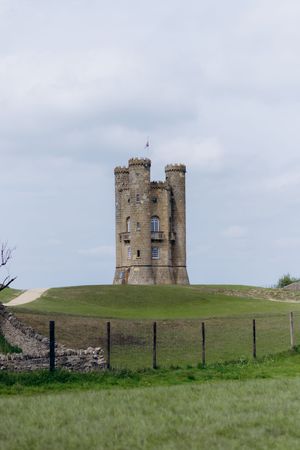 Broadway Tower, a historic folly castle atop a green hill in the Cotswolds, England, under a cloudy sky.