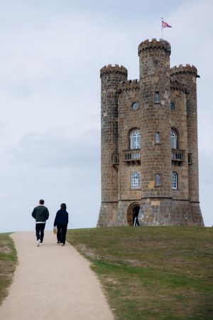 An engaged couple walking along a path toward Broadway Tower, a historic folly castle in the Cotswolds, England.