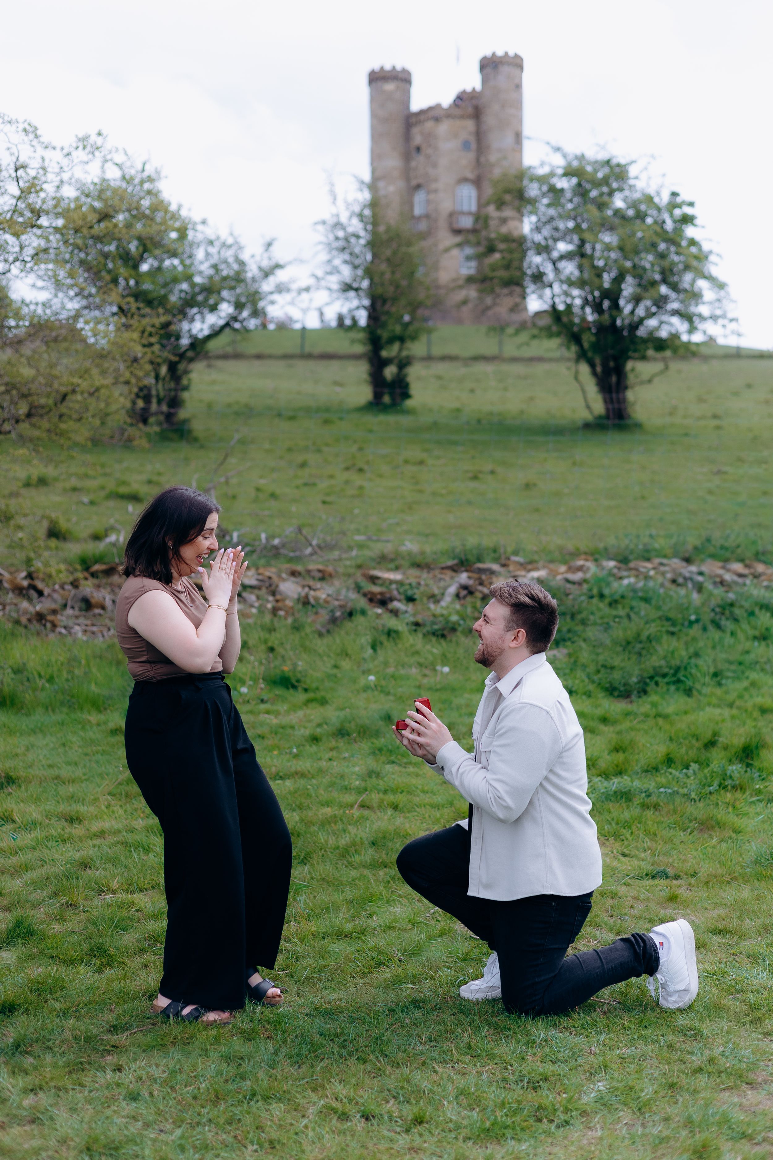 Man proposes on one knee to woman in front of historic Broadway Tower on a green hillside in England.