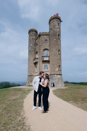 Romantic couple kissing in front of Broadway Tower folly castle under an overcast sky.