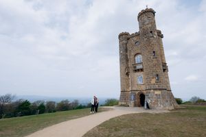 Two people stand beside a historic Broadway Tower on a grassy hill under a cloudy sky.