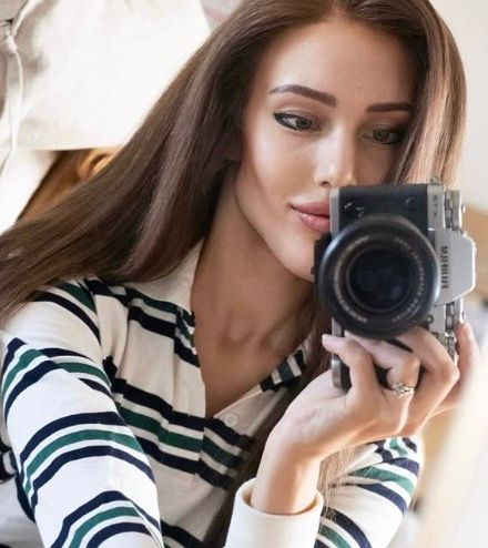 Young woman with long brown hair holding a Fujifilm camera up to take a photo, wearing a striped shirt.