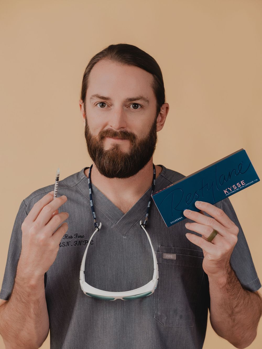 A bearded male medical professional in gray scrubs holds a syringe and a teal Kysse filler box against a beige background.
