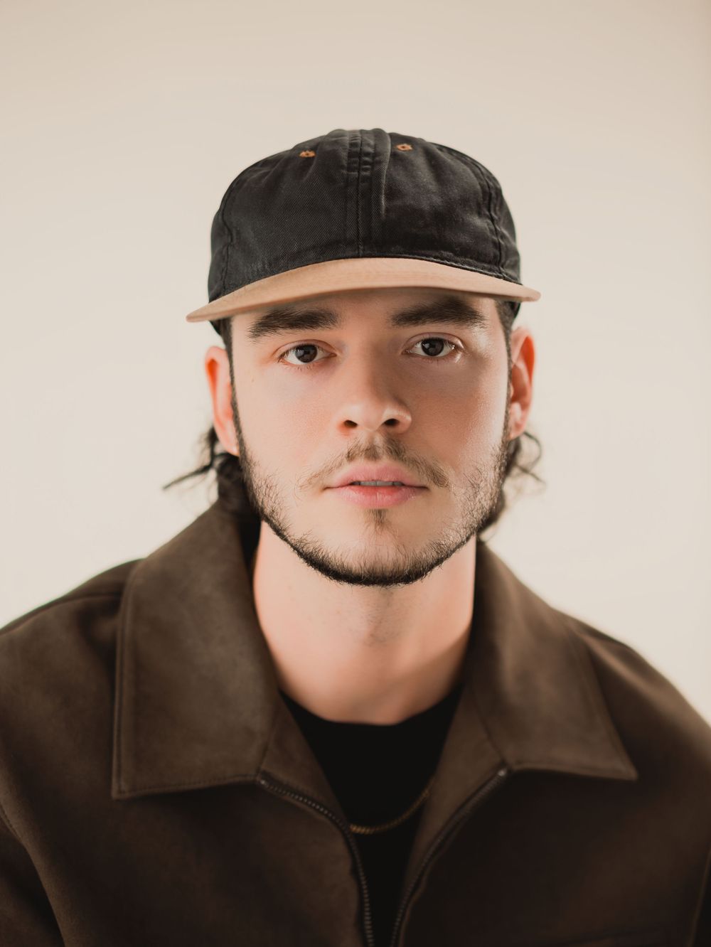 Young man with dark beard wearing a black cap and brown jacket poses for a portrait against a neutral background.