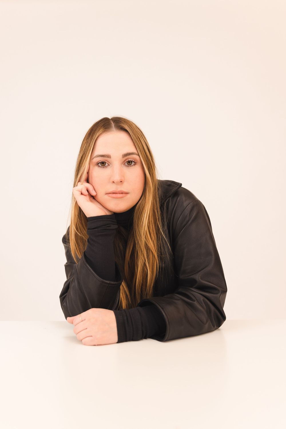 Young woman with long blonde hair in black turtleneck jacket resting chin on hand at white table.