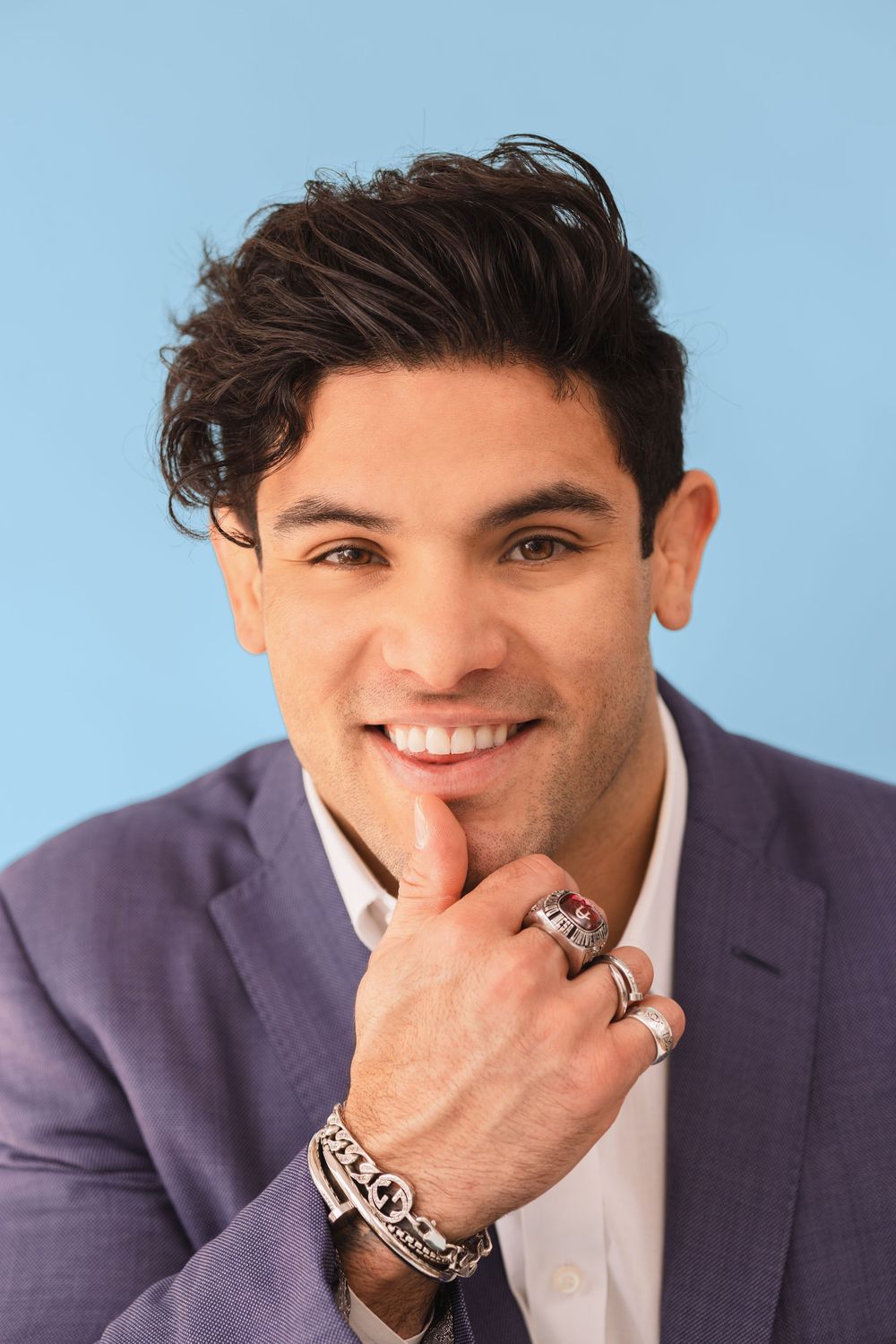 Smiling young man in purple blazer with bracelets and rings, posing confidently against a light blue background.