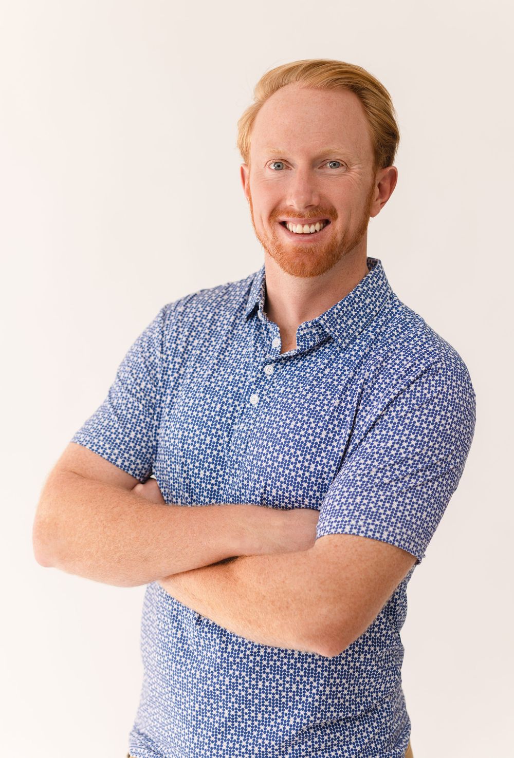 Smiling redhead man with crossed arms wearing a blue patterned polo shirt, professional headshot on white background.