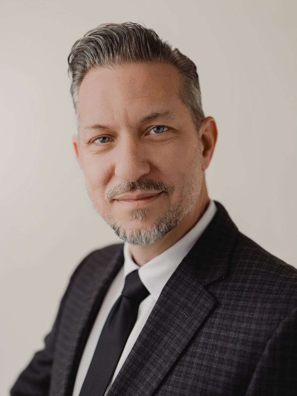 Professional headshot of a middle-aged man with gray hair wearing a dark suit and black tie.