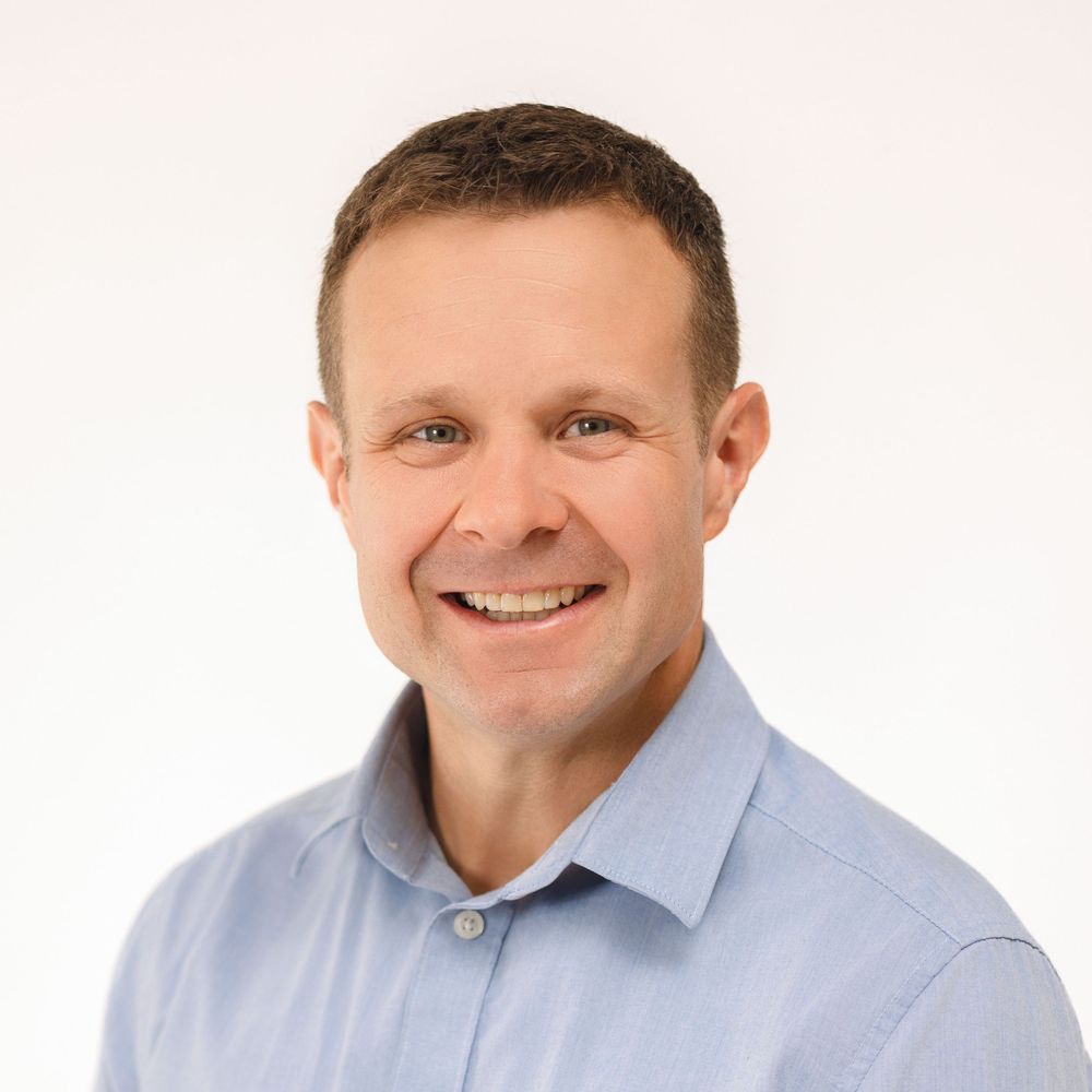 A smiling middle-aged man with short brown hair wearing a light blue shirt against a white background.