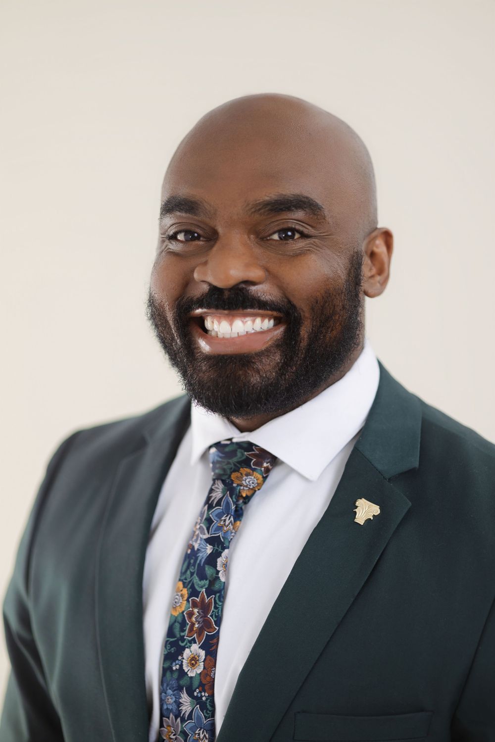 Smiling bald man with beard wearing green suit and floral tie in professional headshot photo.
