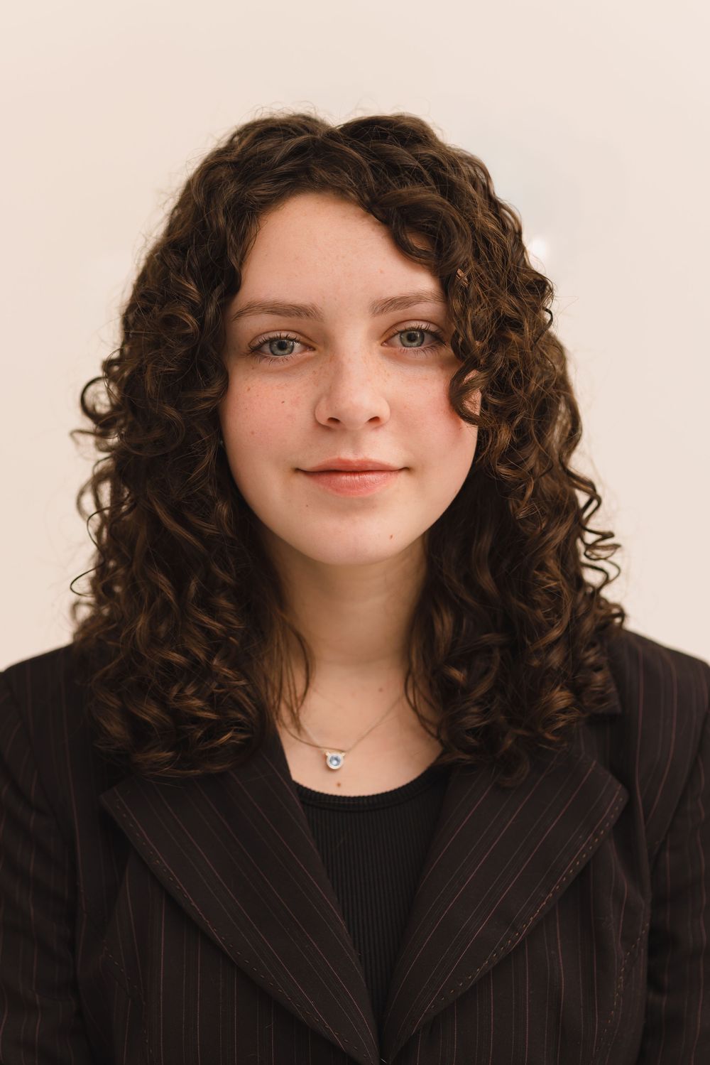 Young woman with curly brown hair wearing a dark blazer and necklace, posing for a professional headshot photo.