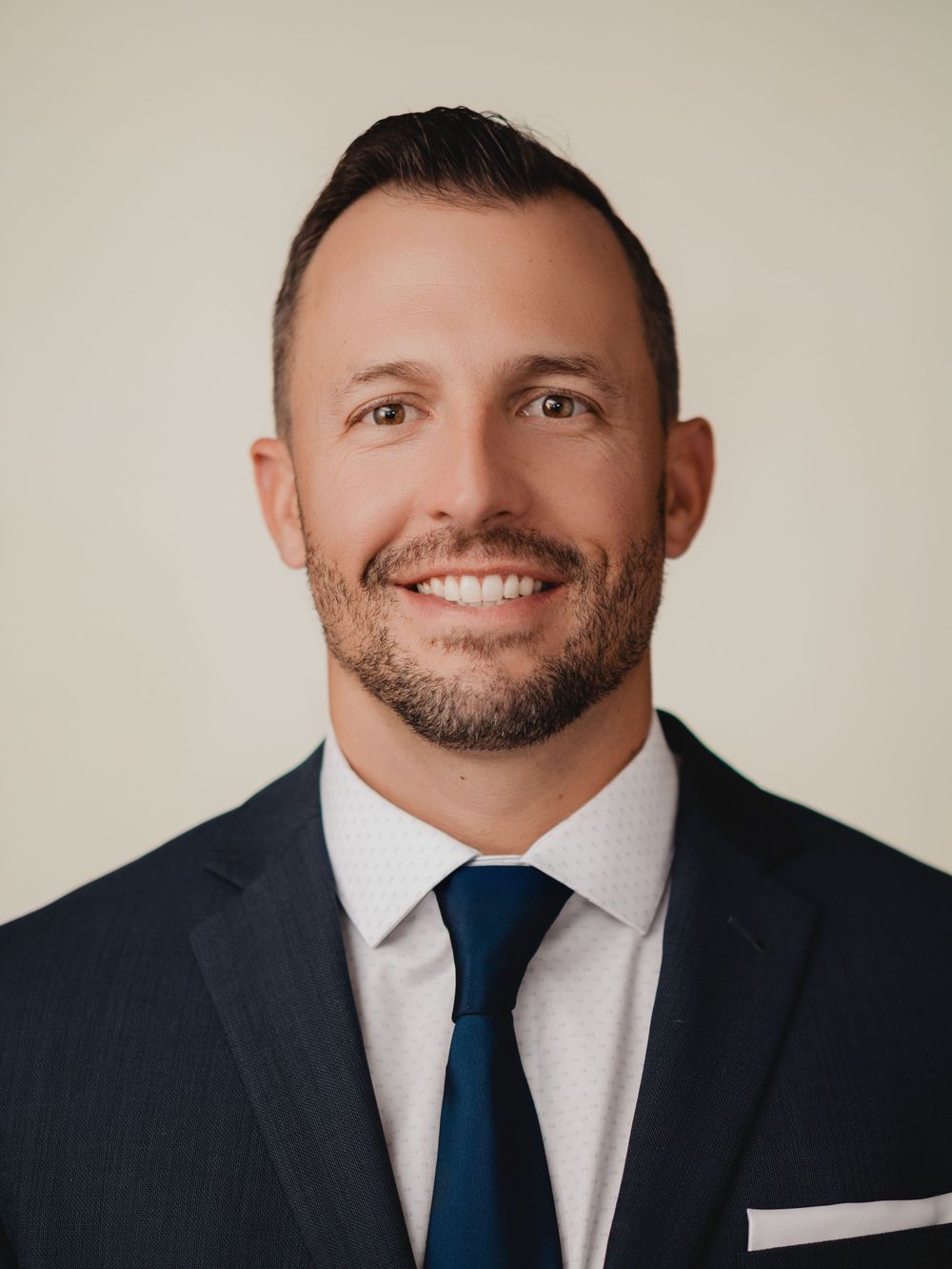 Professional headshot of a smiling man in a navy suit with blue tie and white pocket square.