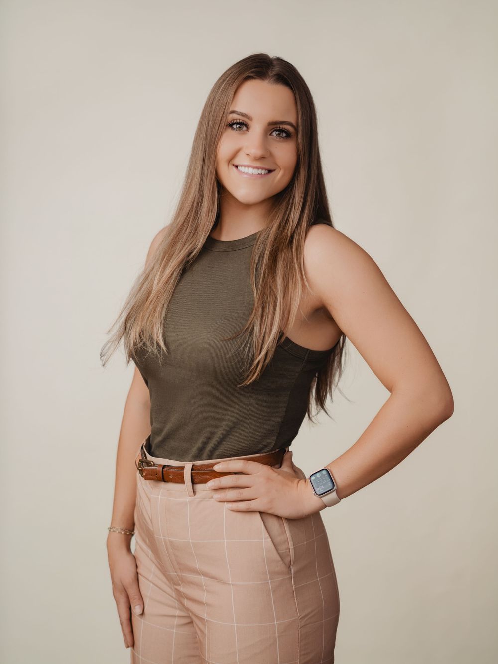 Young woman smiling in olive top and pink plaid pants, posing confidently against a neutral background.