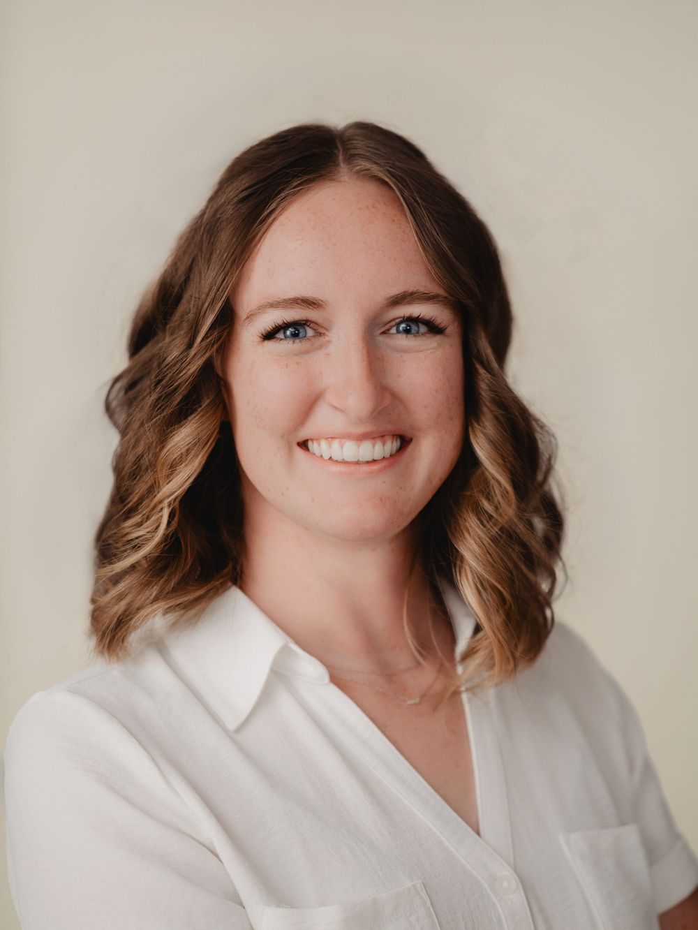Professional headshot of a smiling woman with brown wavy hair wearing a white blouse against a neutral background.