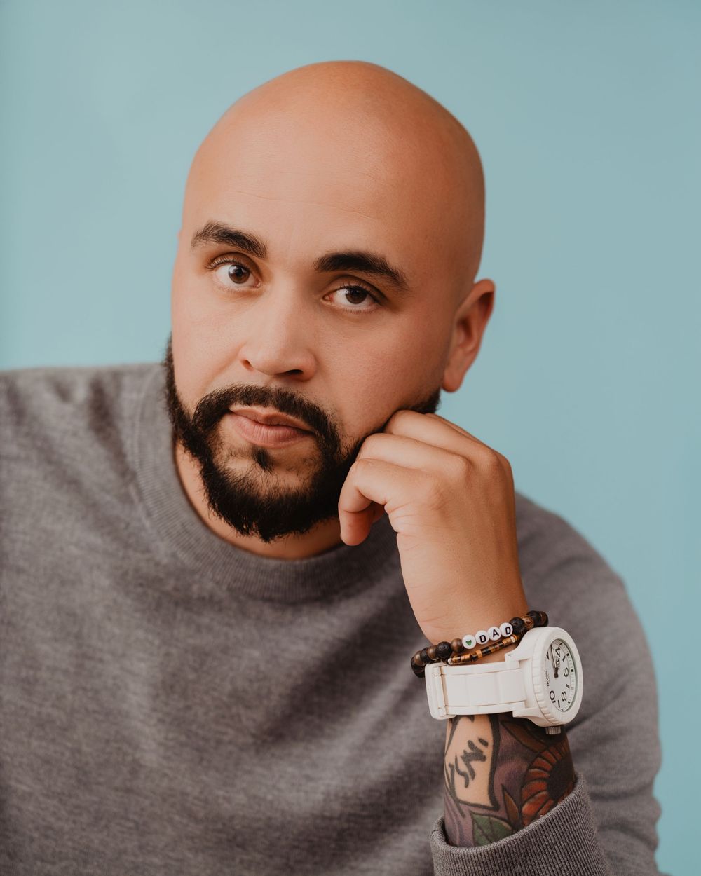 Bald man with beard poses thoughtfully, wearing gray sweater, white watch, and beaded bracelet against blue background.