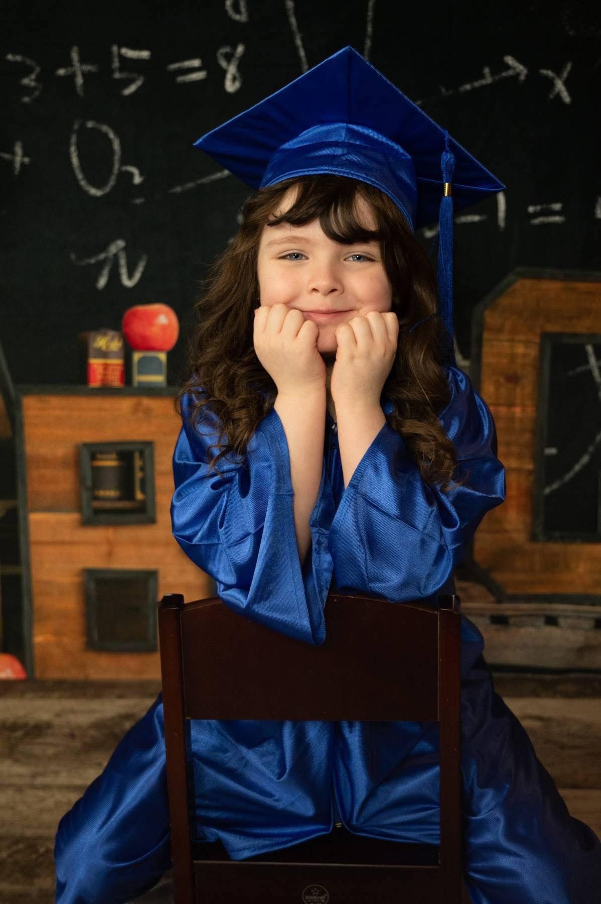 Adorable young girl in blue graduation cap and gown sitting in chair before a chalkboard, smiling.