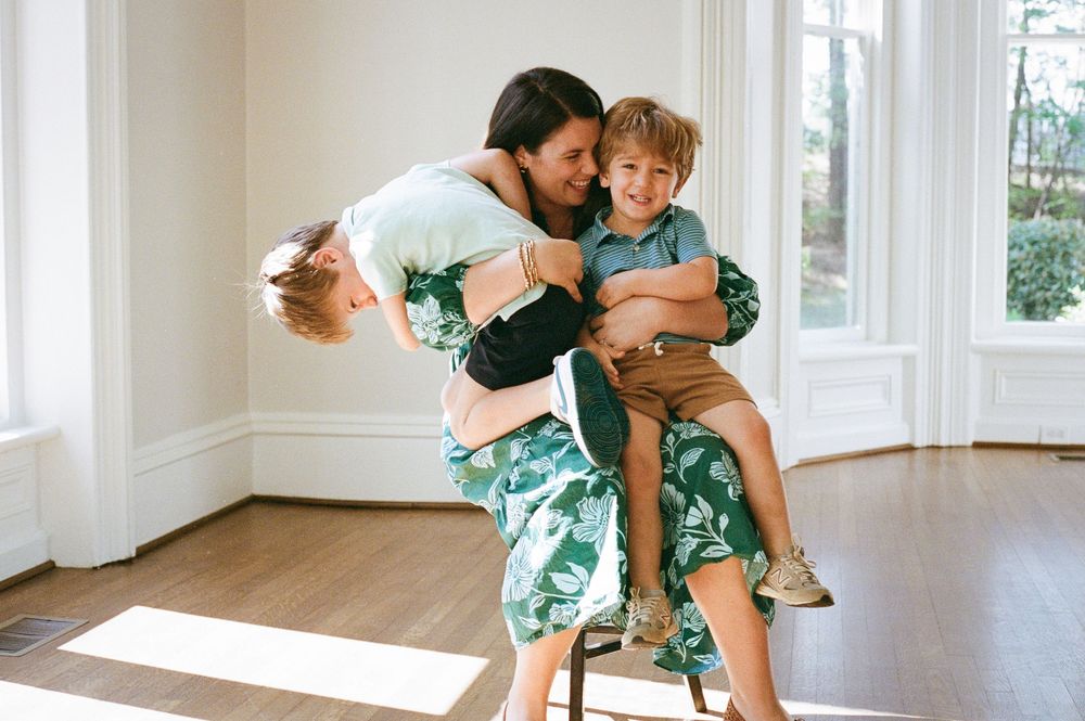 A joyful mother playfully hugs her two children in a bright, sunlit room with hardwood floors.