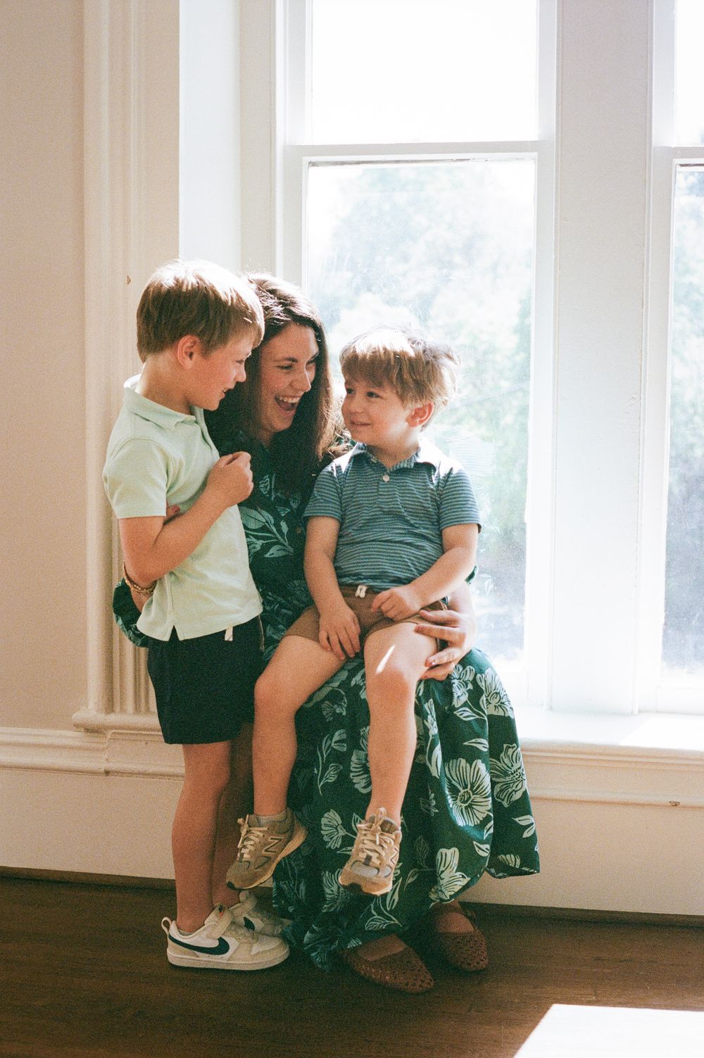 A mother sits by a window laughing with her two young boys in a bright, airy room.
