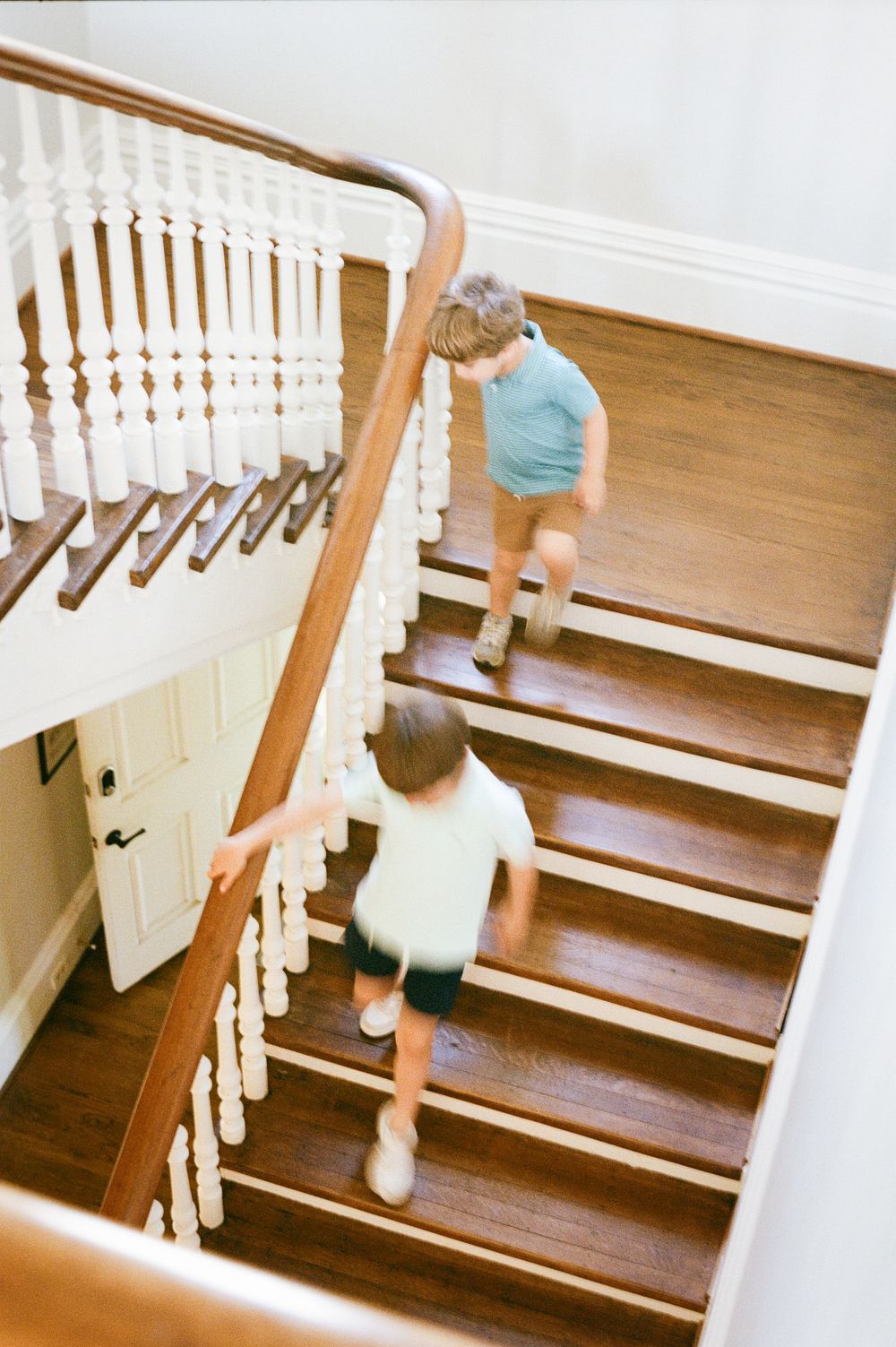 Two children running down wooden stairs with white balusters in a classic home interior.