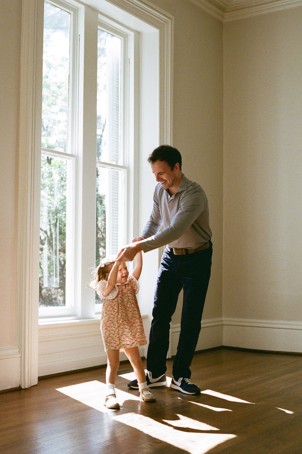 Father dancing with young daughter in sunlit room with hardwood floors and large windows.