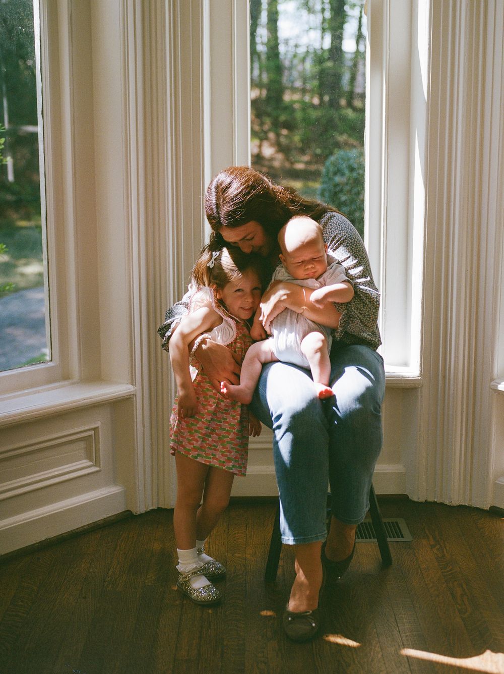 Mother hugging two young children by sunny bay windows in a bright home.