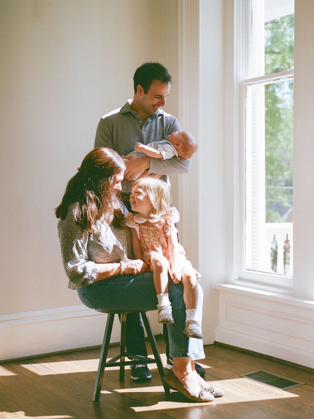 Happy family of four with newborn baby and toddler pose by bright window in airy room.