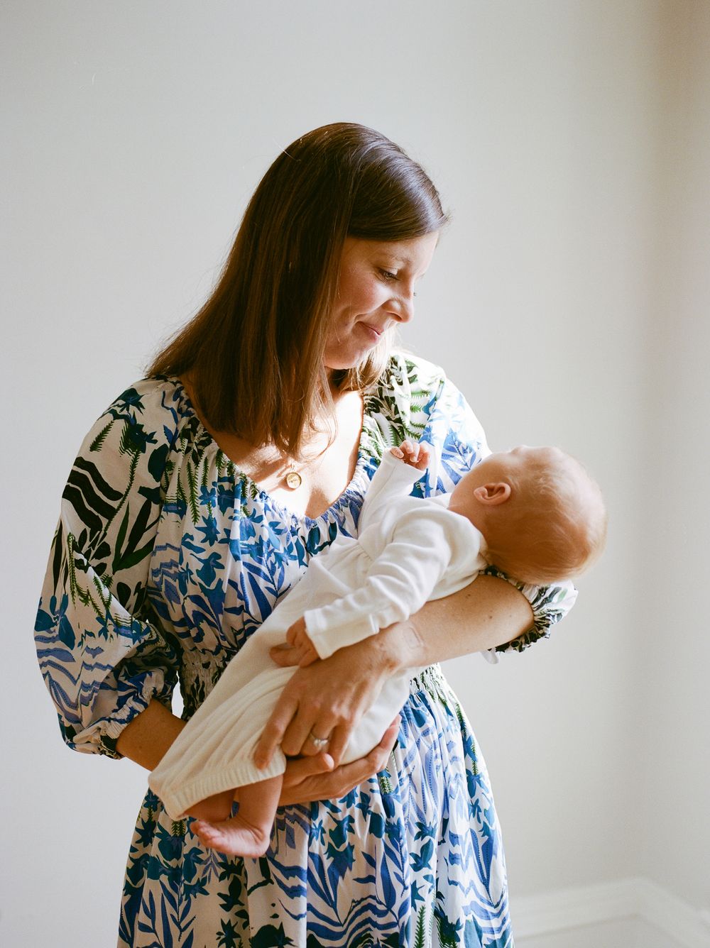 Smiling brunette mother in blue floral dress lovingly gazing at her newborn baby indoors.