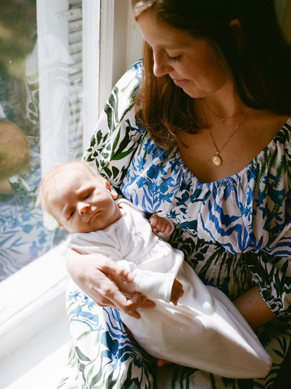 Mother in blue floral dress tenderly holds swaddled newborn baby by sunlit window.