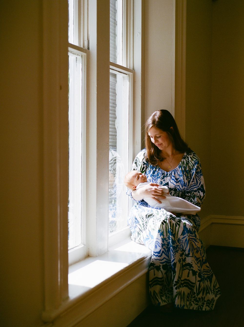 A woman in a blue floral dress sits by a sunlit window, tenderly cradling a newborn baby in her arms.