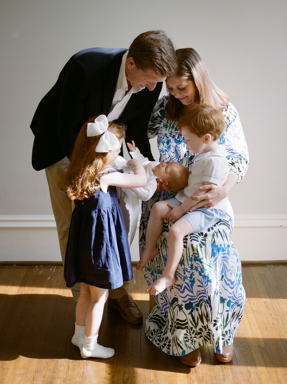 A family of five gathers closely, parents and two children admiring a newborn baby in a warmly lit room.