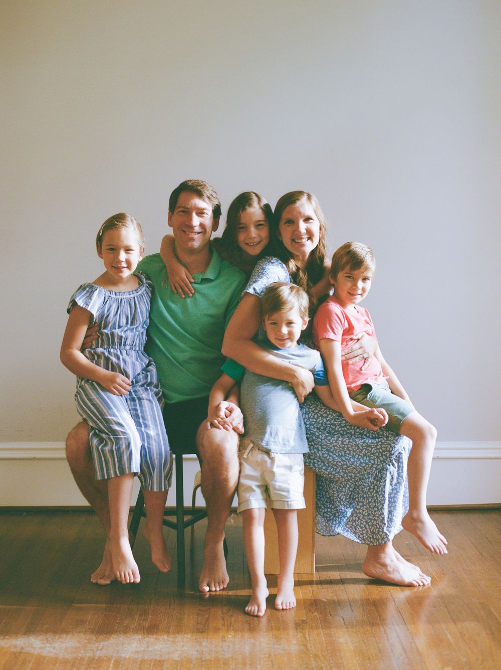 A happy family of six poses together barefoot in a bright indoor setting, smiling warmly for a portrait photo.