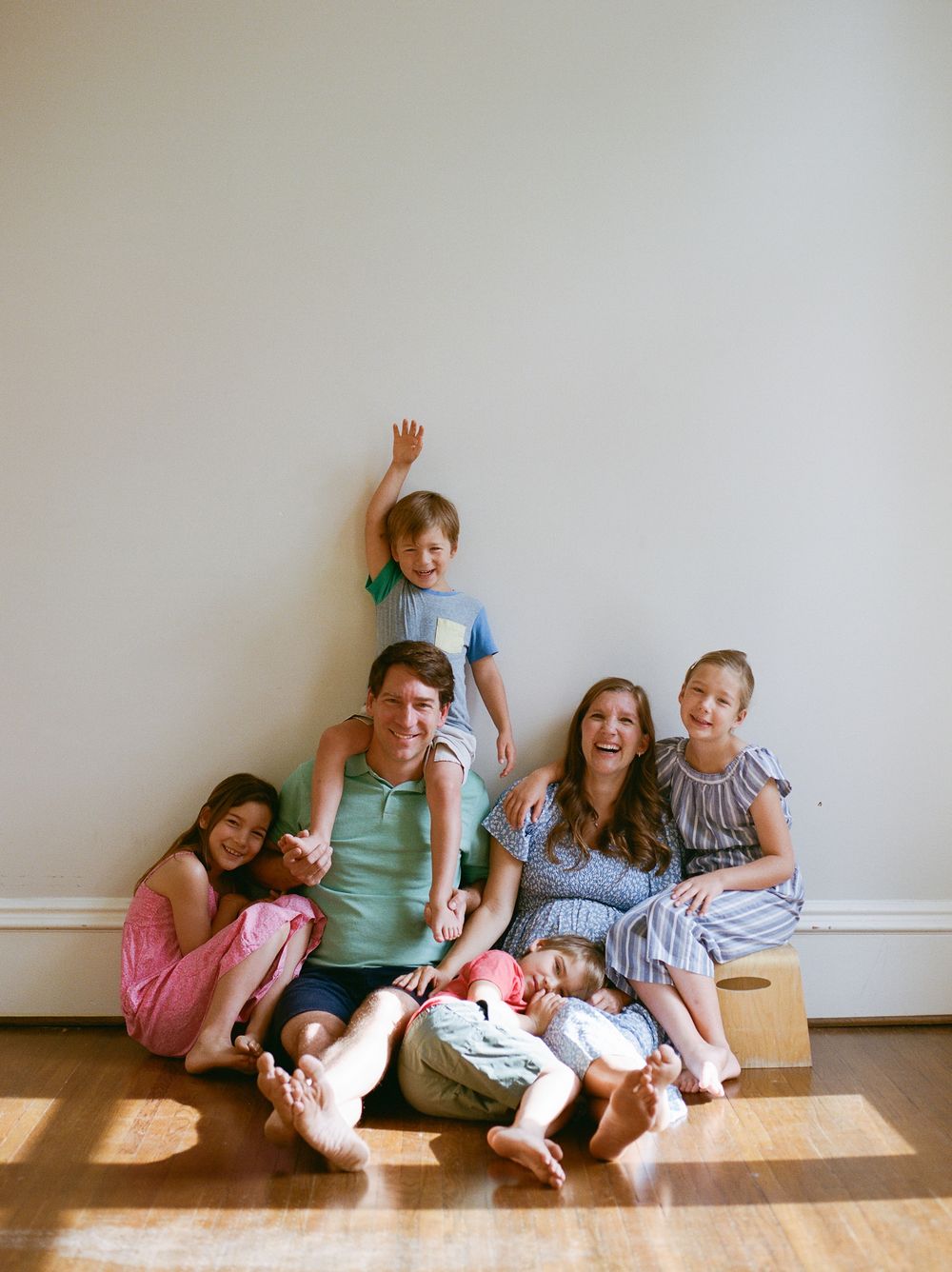 A happy family of six poses casually on a hardwood floor, with two adults and four children smiling and laughing together.