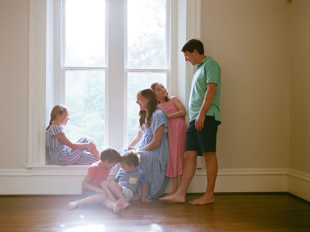 Family gathered by a large window in a bright room, with children playing on the floor and a man standing nearby.