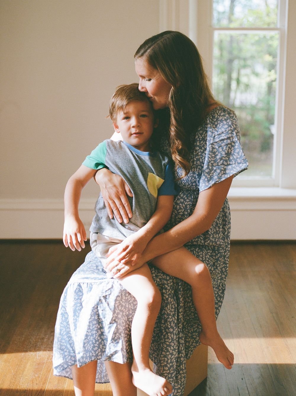 A mother in a floral dress kisses her young son while holding him on her lap near a sunlit window.