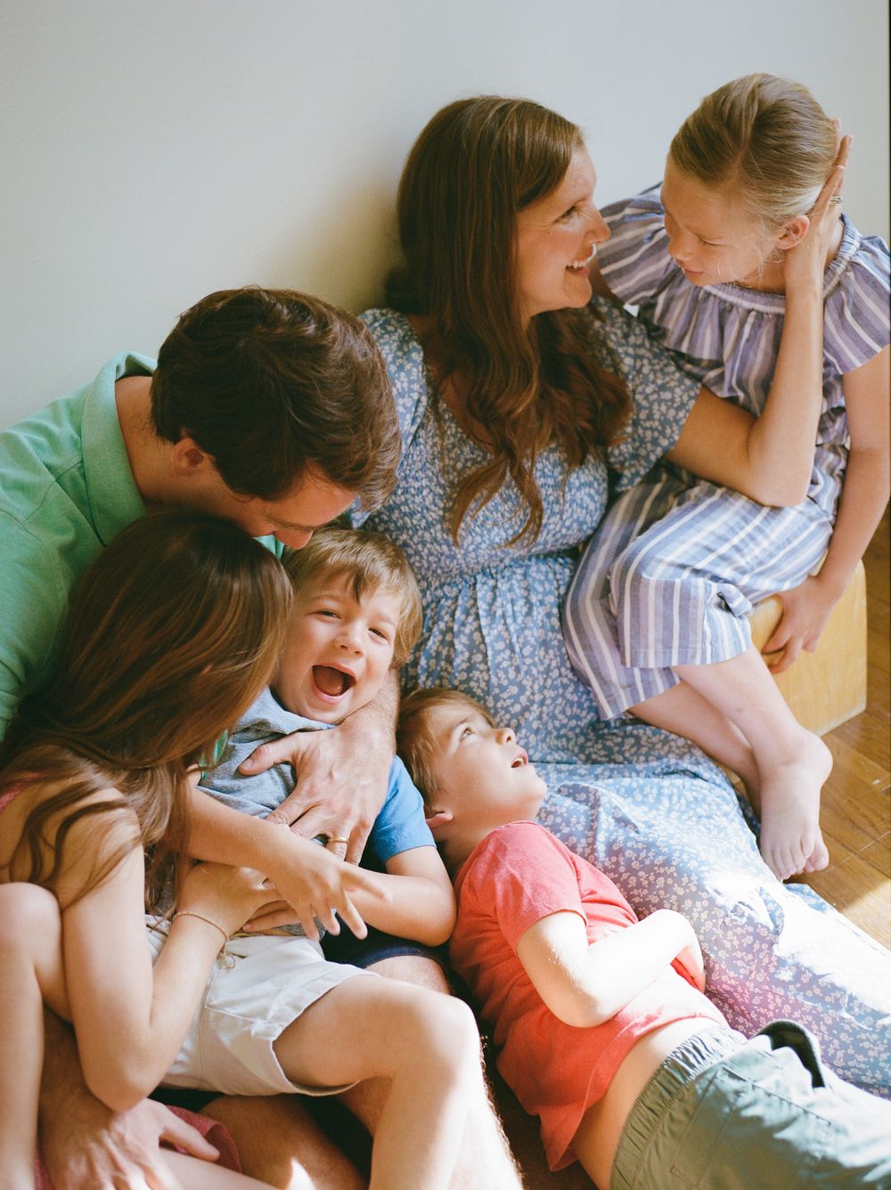 Happy family with four children laughing and cuddling together on a couch in a bright, cozy living room.