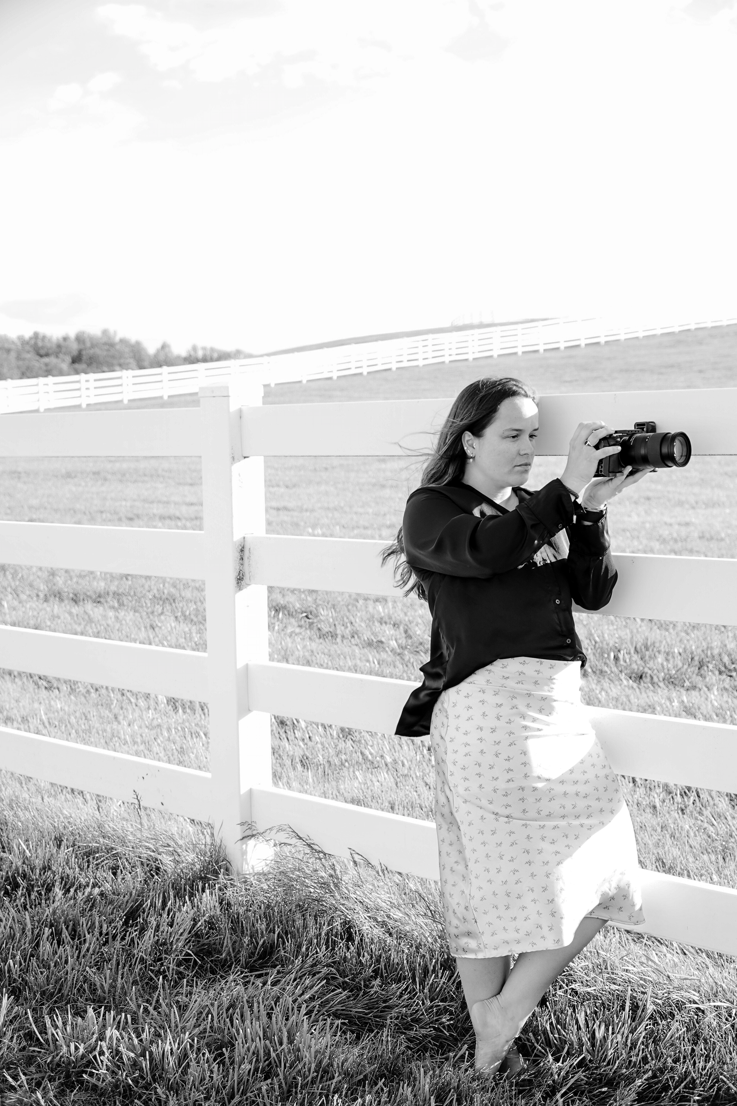 Black and white photo of a woman leaning on a white fence in a field, looking through a camera.