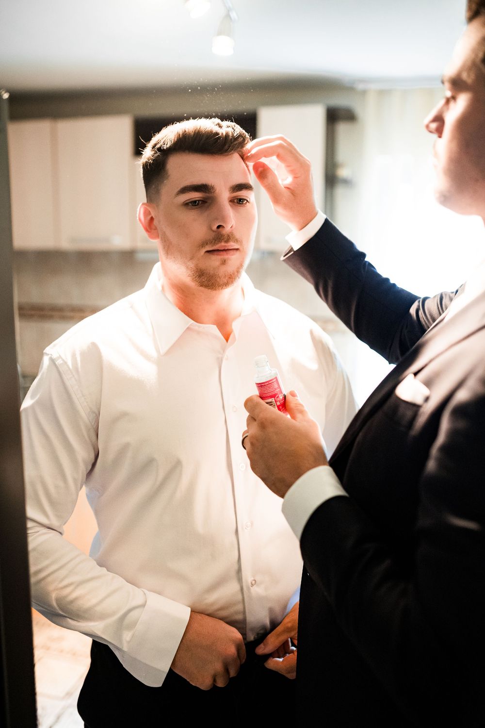 A groomsman helps the groom apply cologne while getting ready for the wedding ceremony.