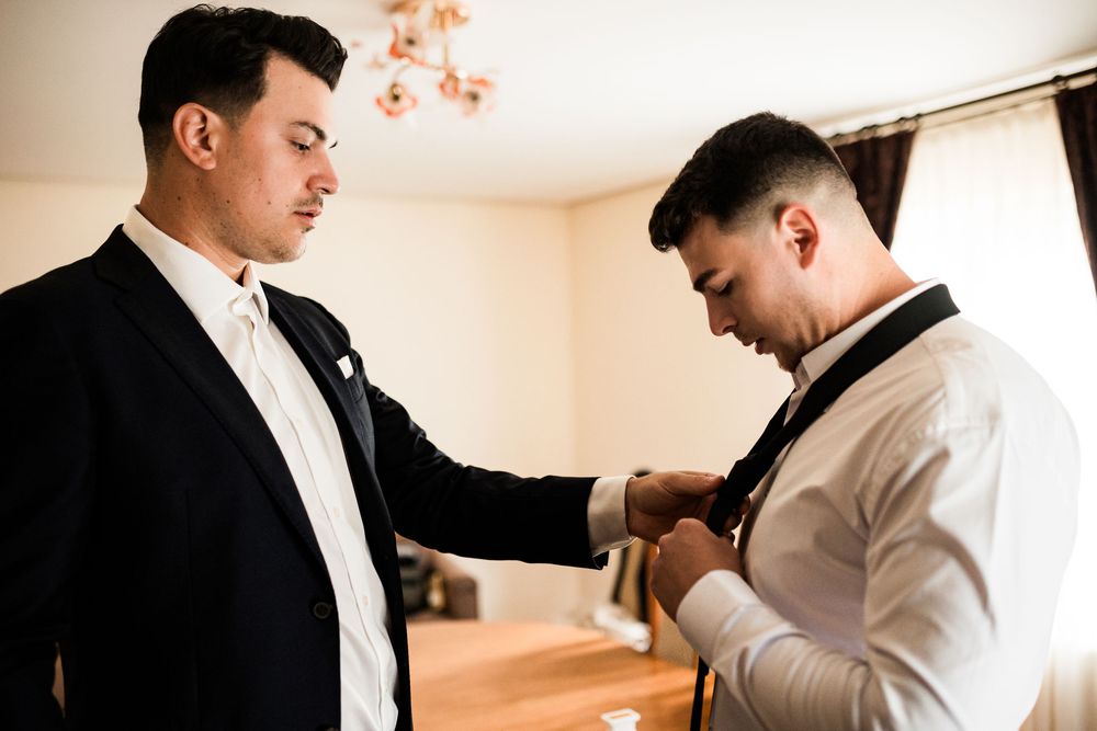 Two men in formal attire help each other prepare for a wedding, adjusting ties and suits.