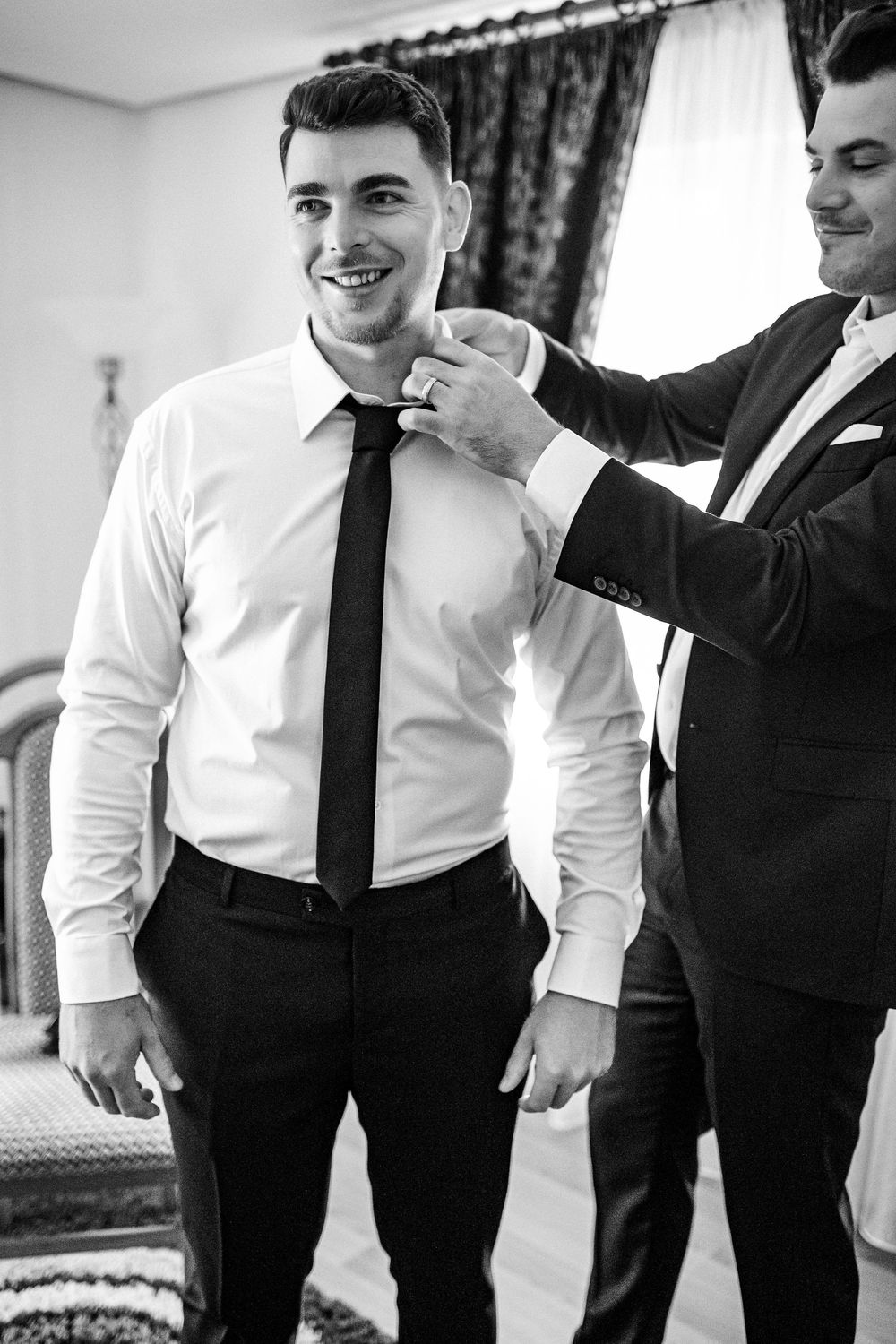 Black and white photo of a smiling groom having his tie adjusted by a groomsman before the wedding.