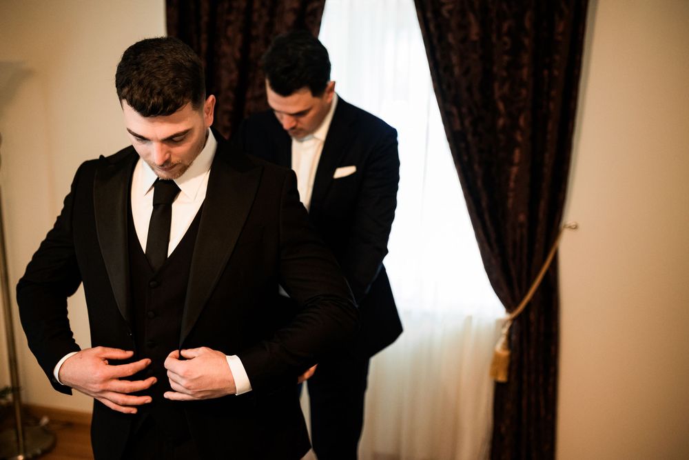 Groom in black tuxedo being helped by groomsman to button jacket before wedding ceremony.
