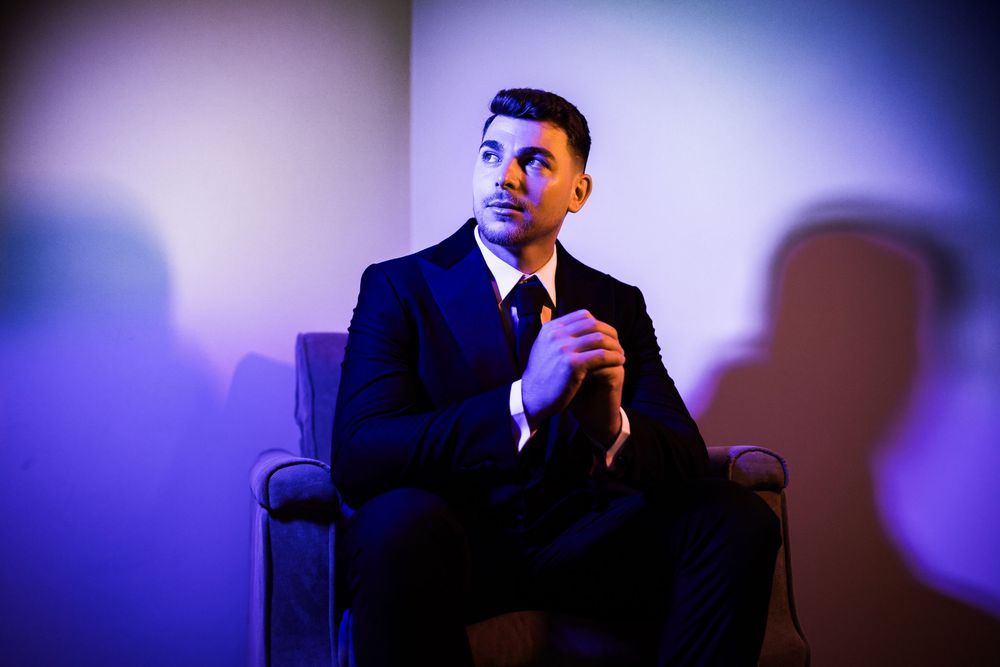 Elegant groom in black suit sitting in chair with dramatic purple and blue lighting backdrop.