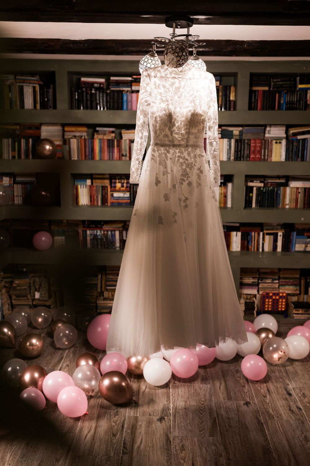 Elegant white lace wedding gown displayed on hanger surrounded by pink and gold balloons in library.