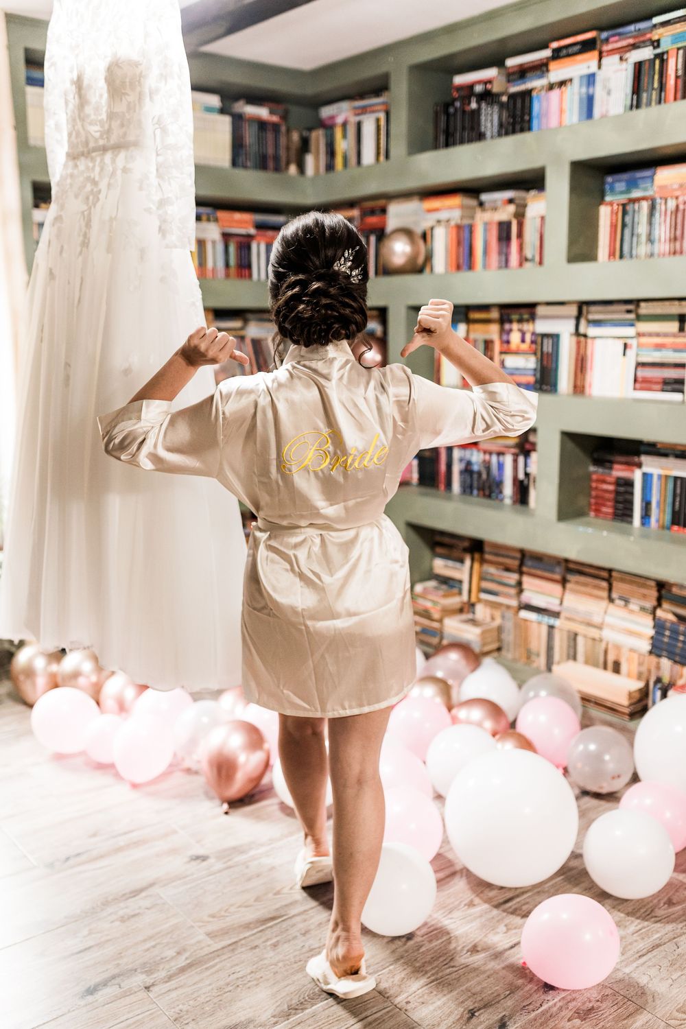 Bride in satin robe flexing arms before her wedding dress in a romantic library setting with balloons.