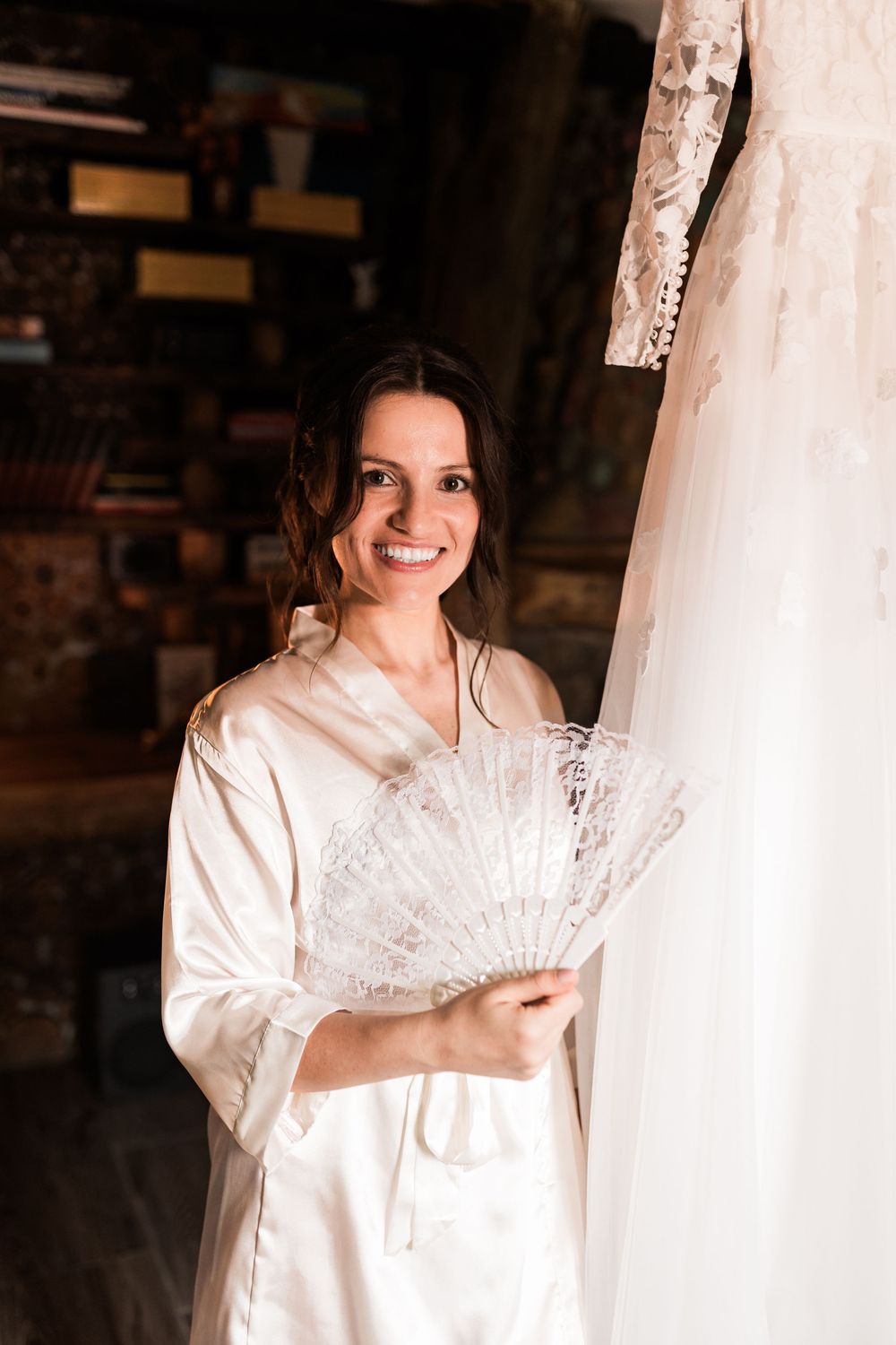 Smiling bride in white satin robe holds a lace fan beside her hanging wedding dress in a warm-lit room.