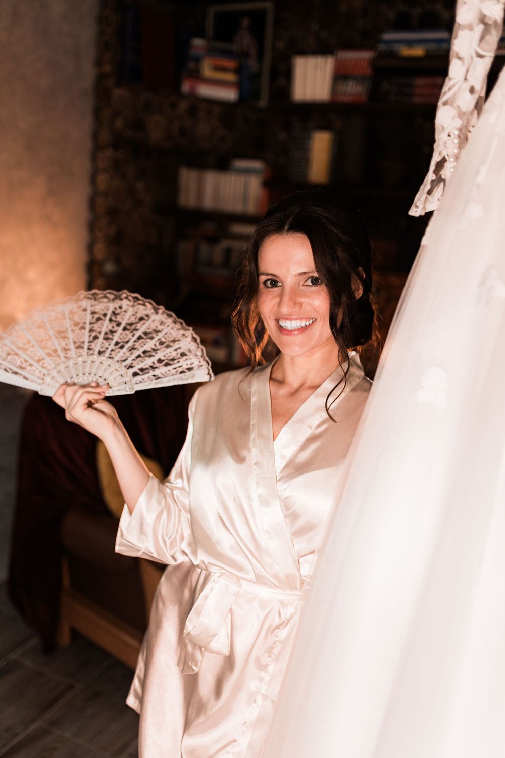 Happy bride in champagne satin robe holds decorative lace fan next to her wedding dress in an elegant room.