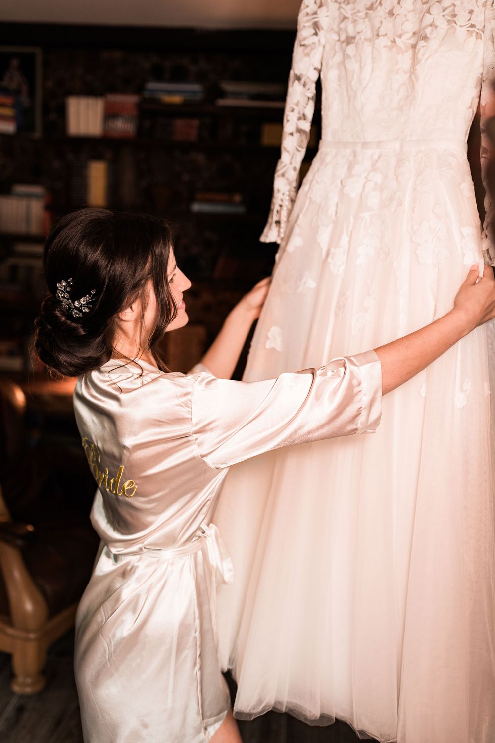 Bride in white satin robe with Bride script reaches up to touch her lace wedding dress hanging in a library.