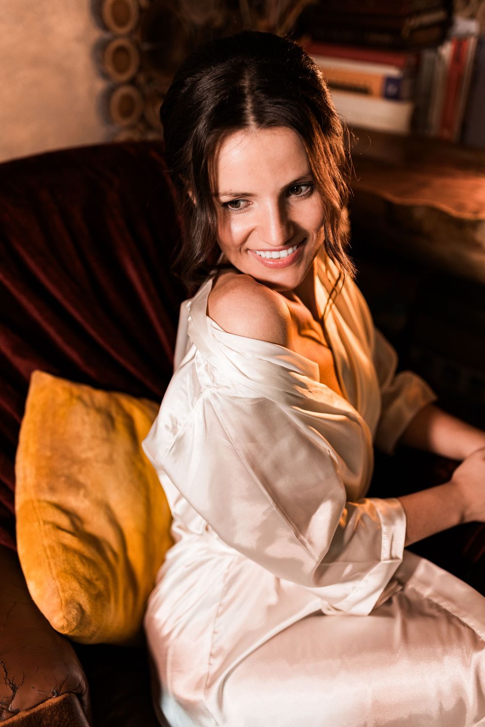 Happy bride in white satin robe sitting on velvet sofa with golden pillow in warm light.