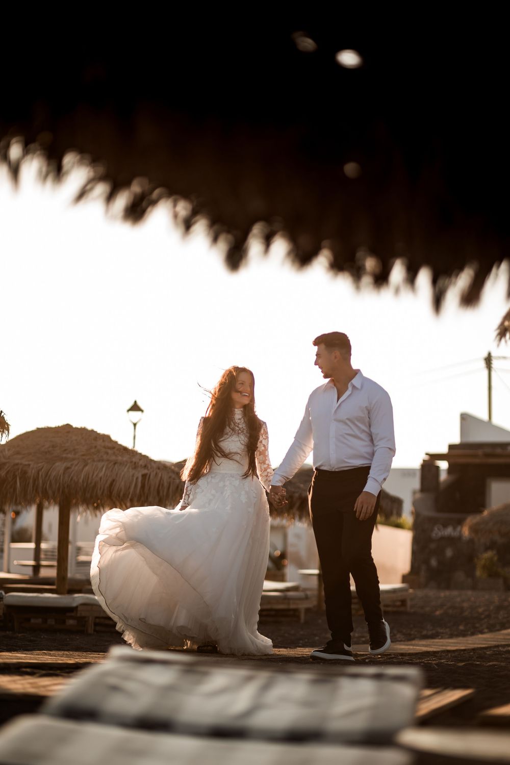 Bride in flowing white gown and groom holding hands at sunset beach venue.
