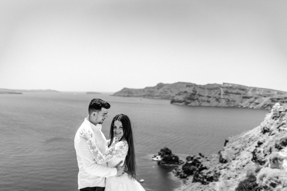 Black and white photo of couple embracing on Santorini cliffside with dramatic sea and island views.